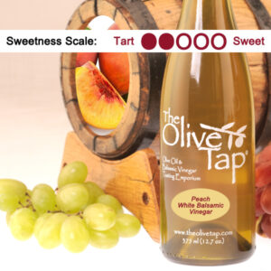 An Olive Tap bottle of Peach White Balsamic Vinegar surrounded by green grapes and wooden barrels.